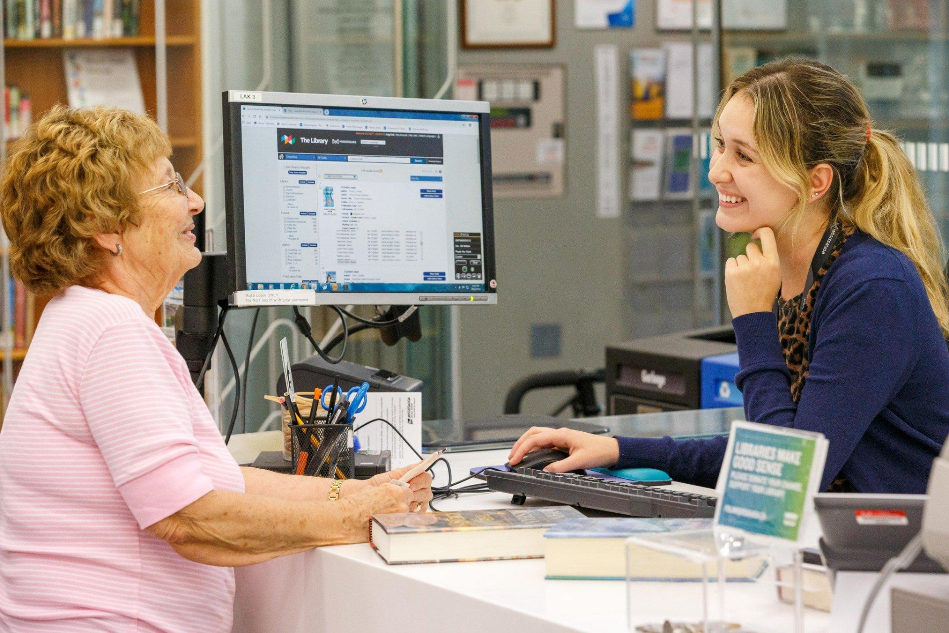 A smiling city library employee helping an older resident at the front desk